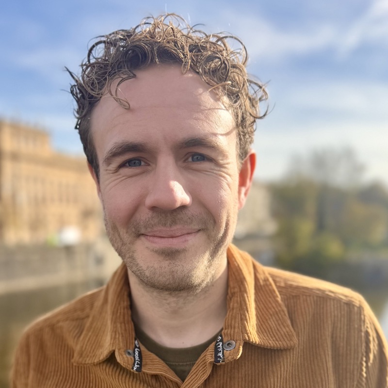 Henry Neves-Charge, smiling, wearing an orange corduroy shirt